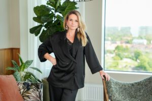 Aros Kapitals nya affärsområdeschef Emelie Tisell poserar på bild med handen på höften framför kontorsmiljö med växter och en grå fåtölj.